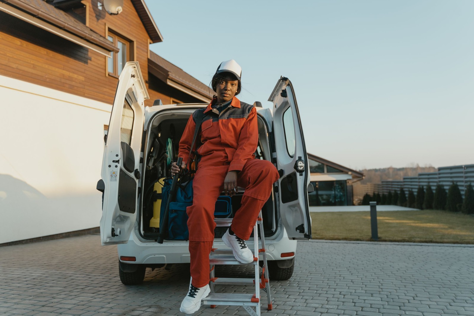 A tradesman in orange overalls sitting on a step ladder at the open back doors of his van.