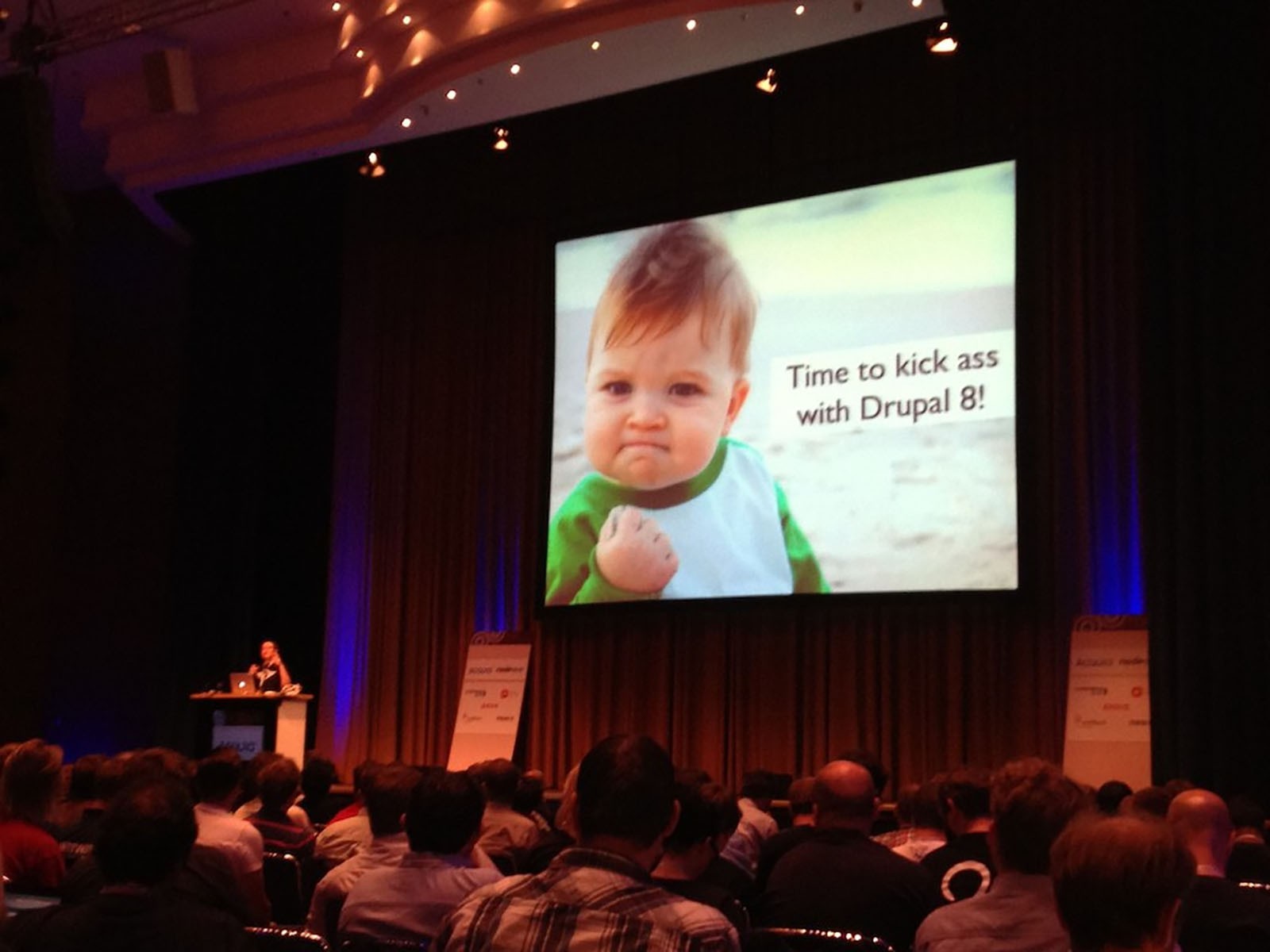 A humorous keynote slide from a DrupalCon. The slide shows a toddler clenching a fist and some wording saying 'Time to kick ass with Drupal 8'.