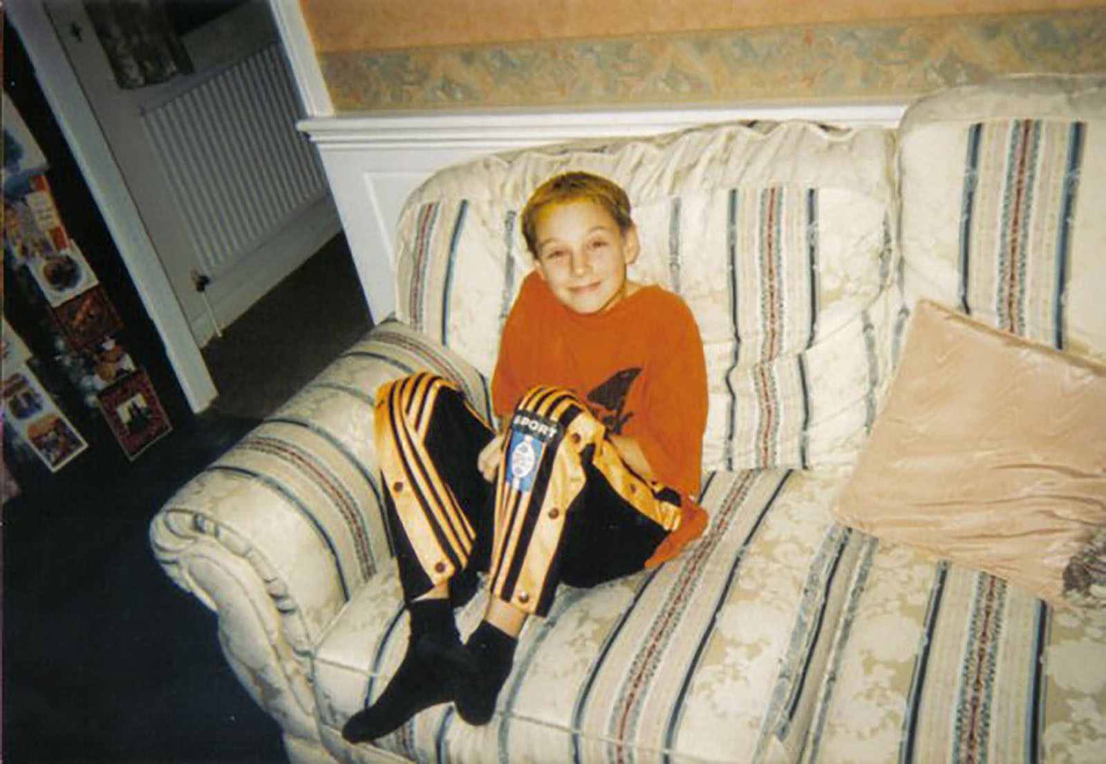 Darren as a young man (circa 10 years old) sat on a white sofa with stripes, wearing an orange t-shirt and black and orange tracksuit bottoms.