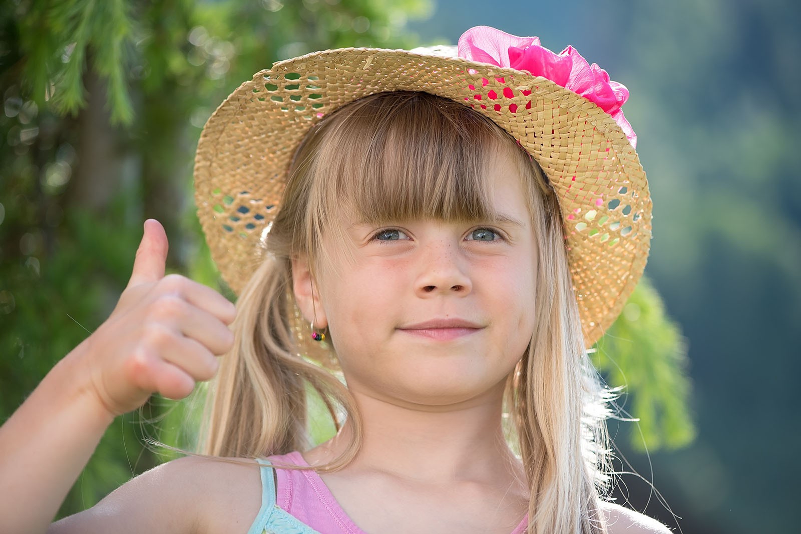 A young child with blonde hair and a summer hat giving a blank expression thumbs up to the camera.