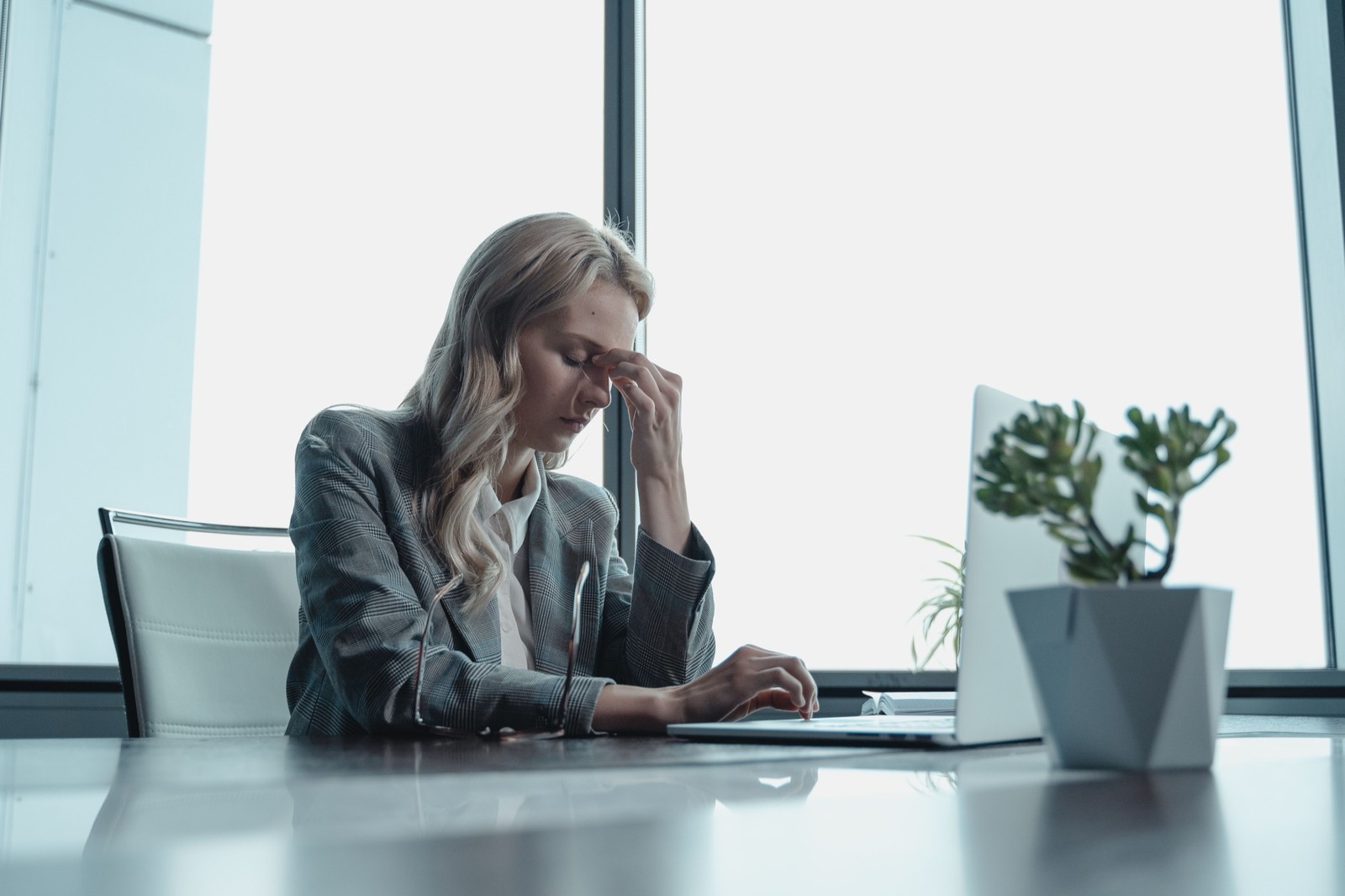 A woman at an office desk pinching the bridge of her nose in front of a laptop, looking stressed.