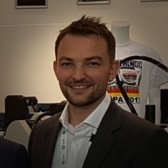 Luke Smith smiling, with short spiky hair and a short beard, wearing a dark jacket and an open-collared shirt, next to printing machinery.