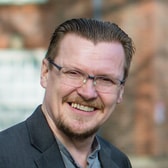 Peter Forest smiling broadly, with slicked-back hair, a moustache and goatee and thin rimless glasses, wearing a dark jacket.