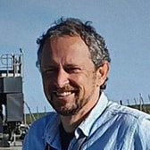 Andy Schofield smiling, with curly hair and stubble, wearing an open-collared denim shirt, outdoors beside industrial equipment.