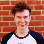Thomas Hiller smiling broadly, with short wavy hair, wearing a baseball-style T-shirt and a cord necklace, in front of a brick wall.