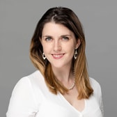 Aoife Breslin smiling, with long hair parted in the middle, wearing drop earrings, a pearl necklace and a light top.
