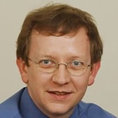 Lawrence Dack with a slight smile, side-parted hair and thin wire-framed glasses, wearing a collared shirt.