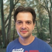 Alexander Benjamin with a slight smile and short swept-back hair, wearing a printed T-shirt, in front of trees.