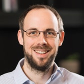 Barry Fisher smiling broadly, with short receding hair, a short beard and thin rectangular glasses, wearing a pinstriped collared shirt.