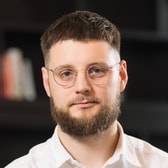 Darren Fisher with a calm, closed-mouth smile, short cropped hair, a full beard and round wire-framed glasses, wearing a white open-collared shirt.