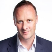 John Kerr with a slight smile and short receding hair, wearing a dark blazer over a striped open-collared shirt.