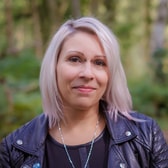 Julia Krajewska with a gentle smile and shoulder-length light hair, wearing a leather jacket and a fine necklace, in front of woodland.