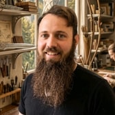 Kristian Johnson smiling, with a long full beard and swept-back hair, wearing a dark T-shirt, in a workshop lined with tools and shelves.