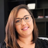 Pri Scarabelli smiling, with shoulder-length straight hair swept to one side and dark cat-eye glasses, wearing a dark top and a fine necklace.