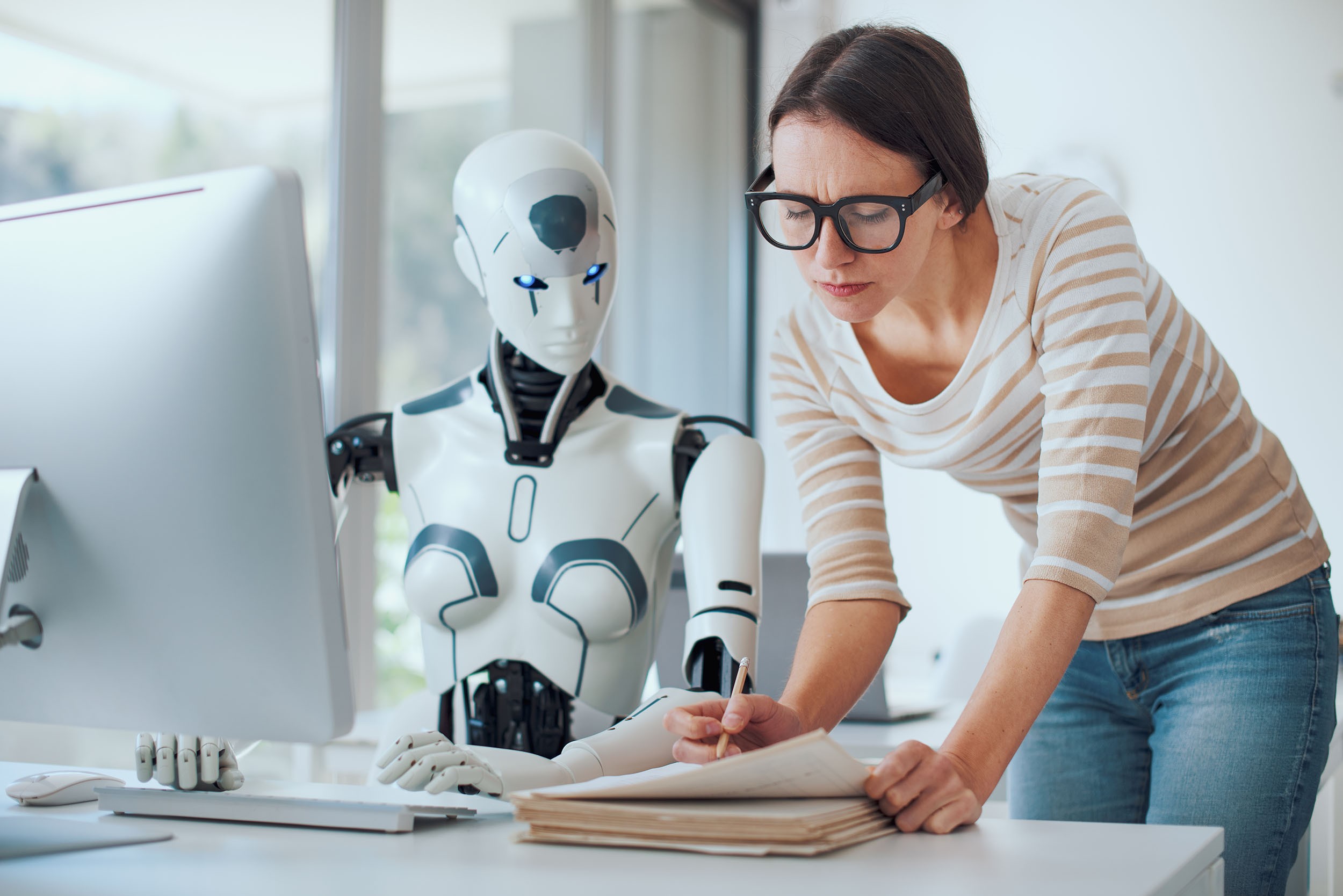A robot at an office desk whose work is being reviewed by a female colleague who looks less than impressed.
