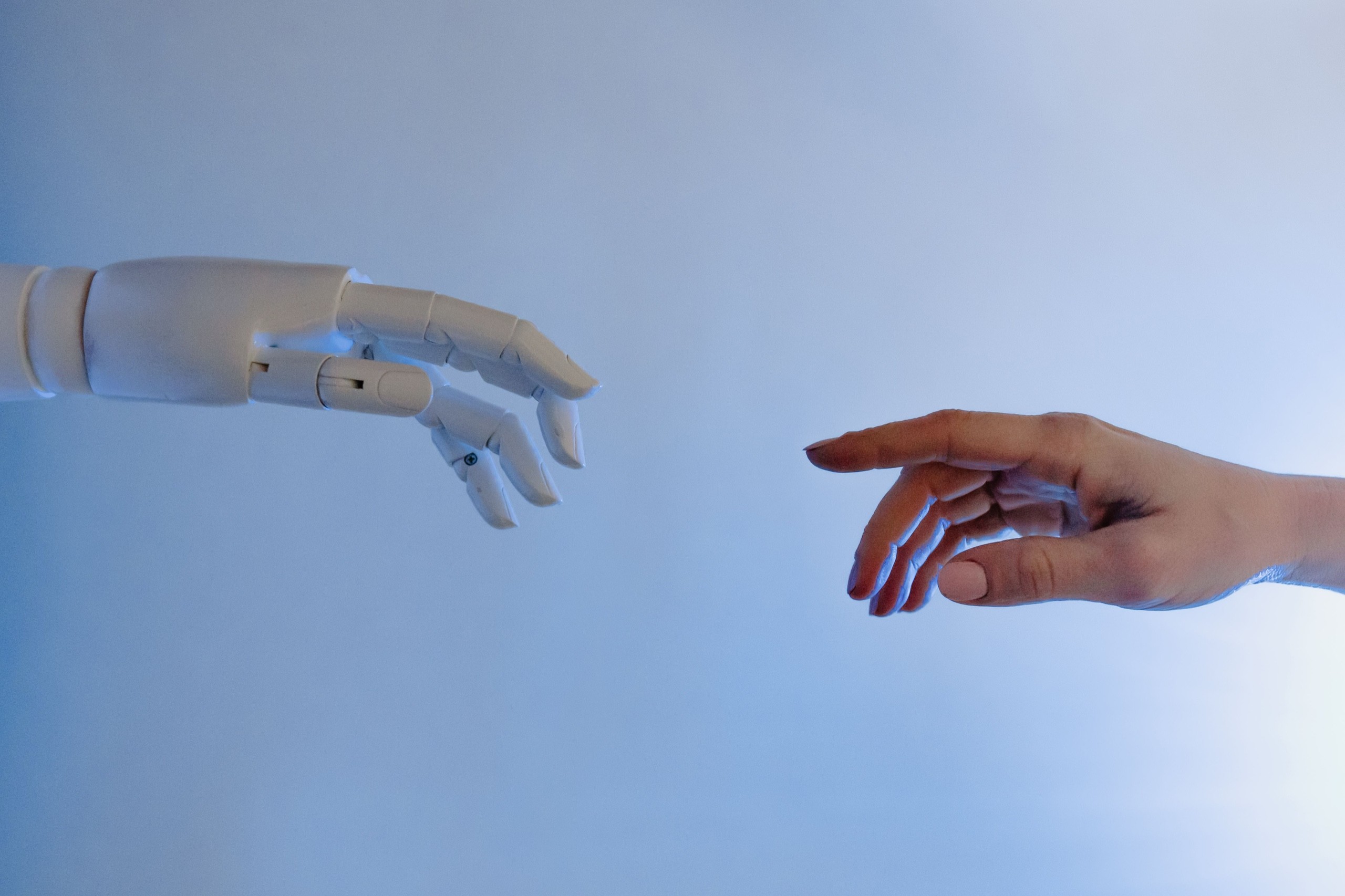 A white robot hand reaching towards a human hand against a pale blue background.