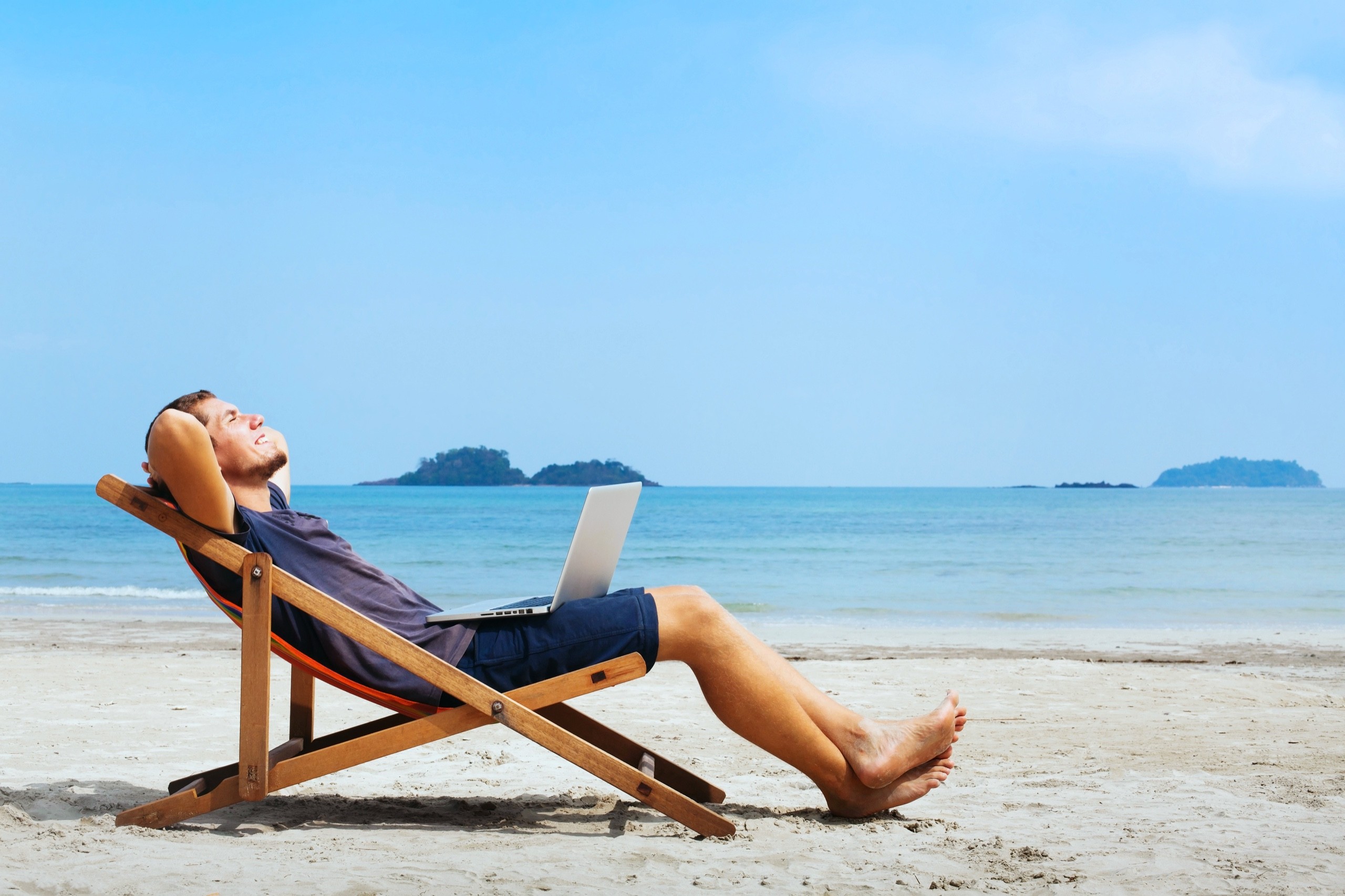 A man in shorts and t-shirt lying back in a deckchair on a white sandy beach with a laptop on his lap.