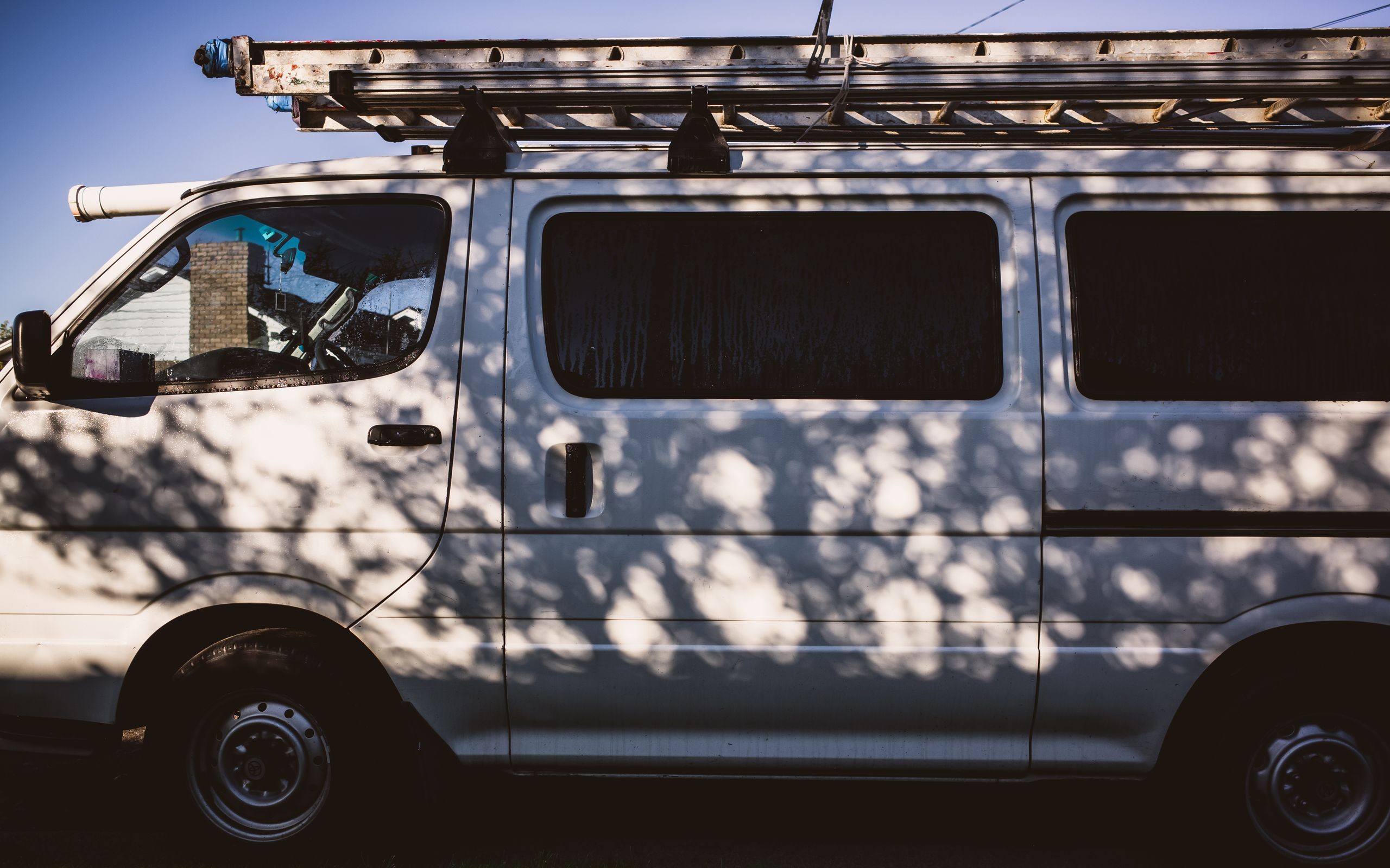A white van with ladders on its roof rack, parked in the sun with tree shadows across its side.