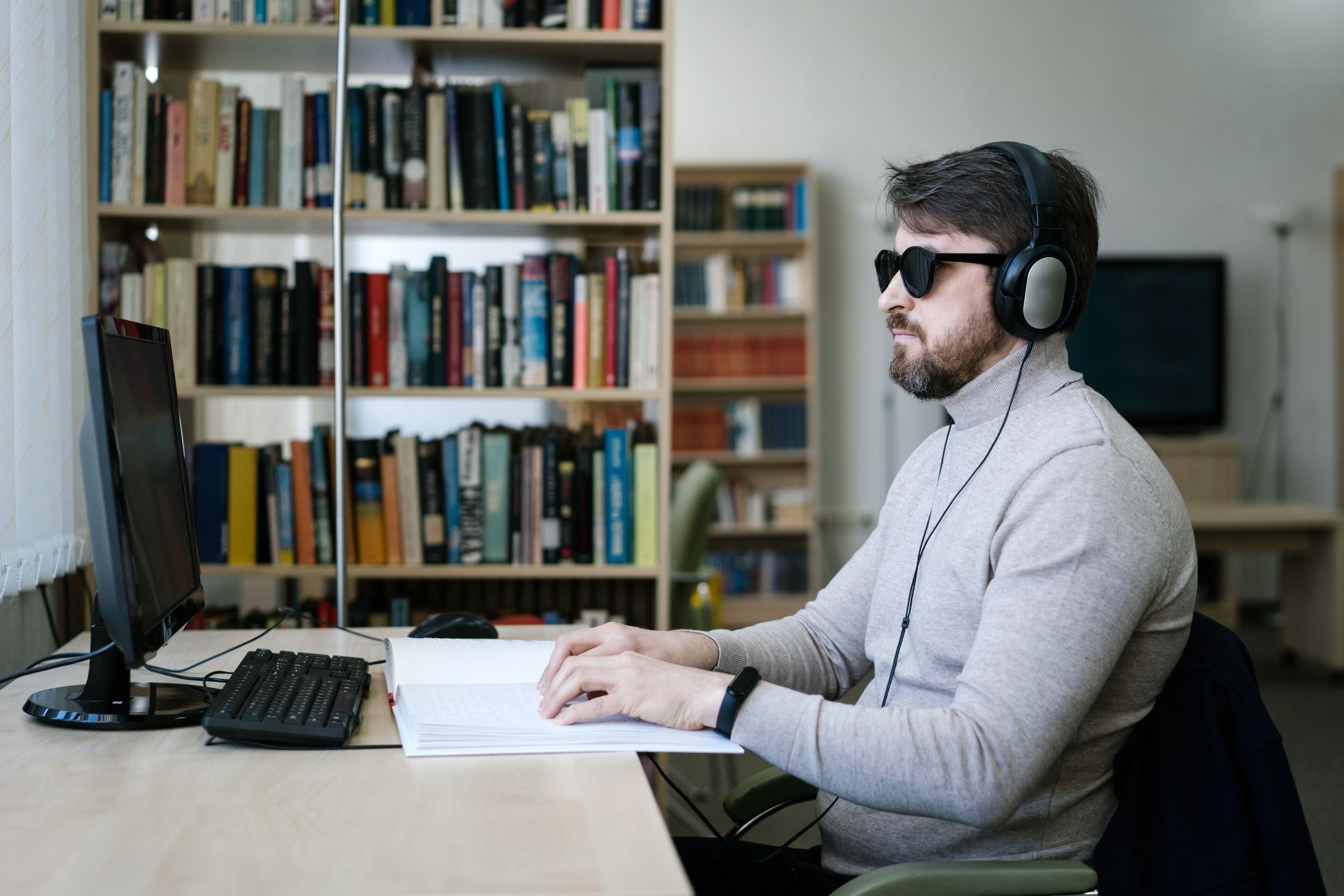 A man wearing dark glasses and headphones reads a Braille book while working at a computer in a library.