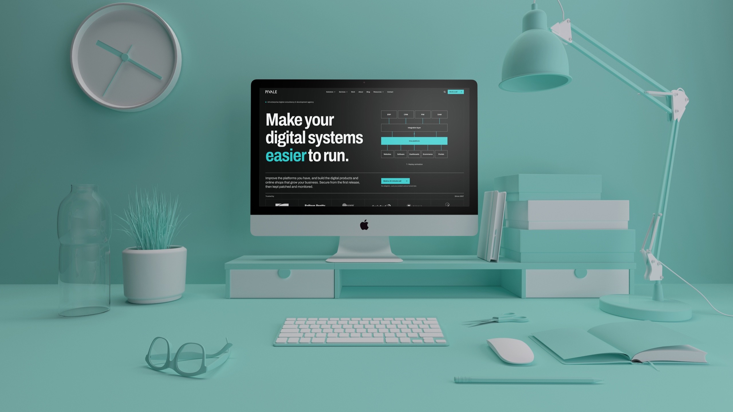 The new Pivale homepage on an iMac, on a teal desk with a clock, lamp, plant and books.