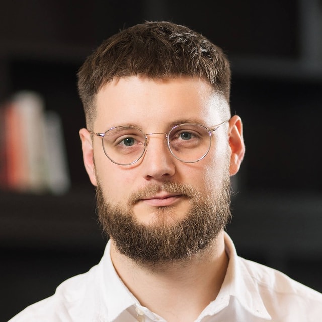 Darren Fisher with a calm, closed-mouth smile, short cropped hair, a full beard and round wire-framed glasses, wearing a white open-collared shirt.