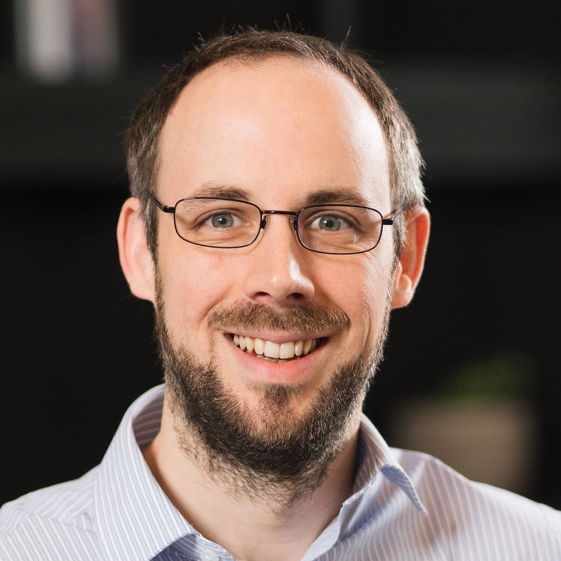Barry Fisher smiling broadly, with short receding hair, a short beard and thin rectangular glasses, wearing a pinstriped collared shirt.